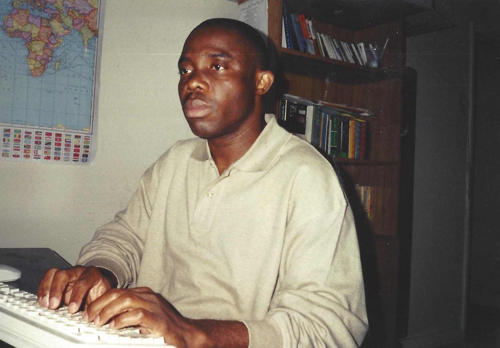 A person wearing a cream-colored shirt types at a computer with a map showing the continent of Africa and a bookcase in the background.