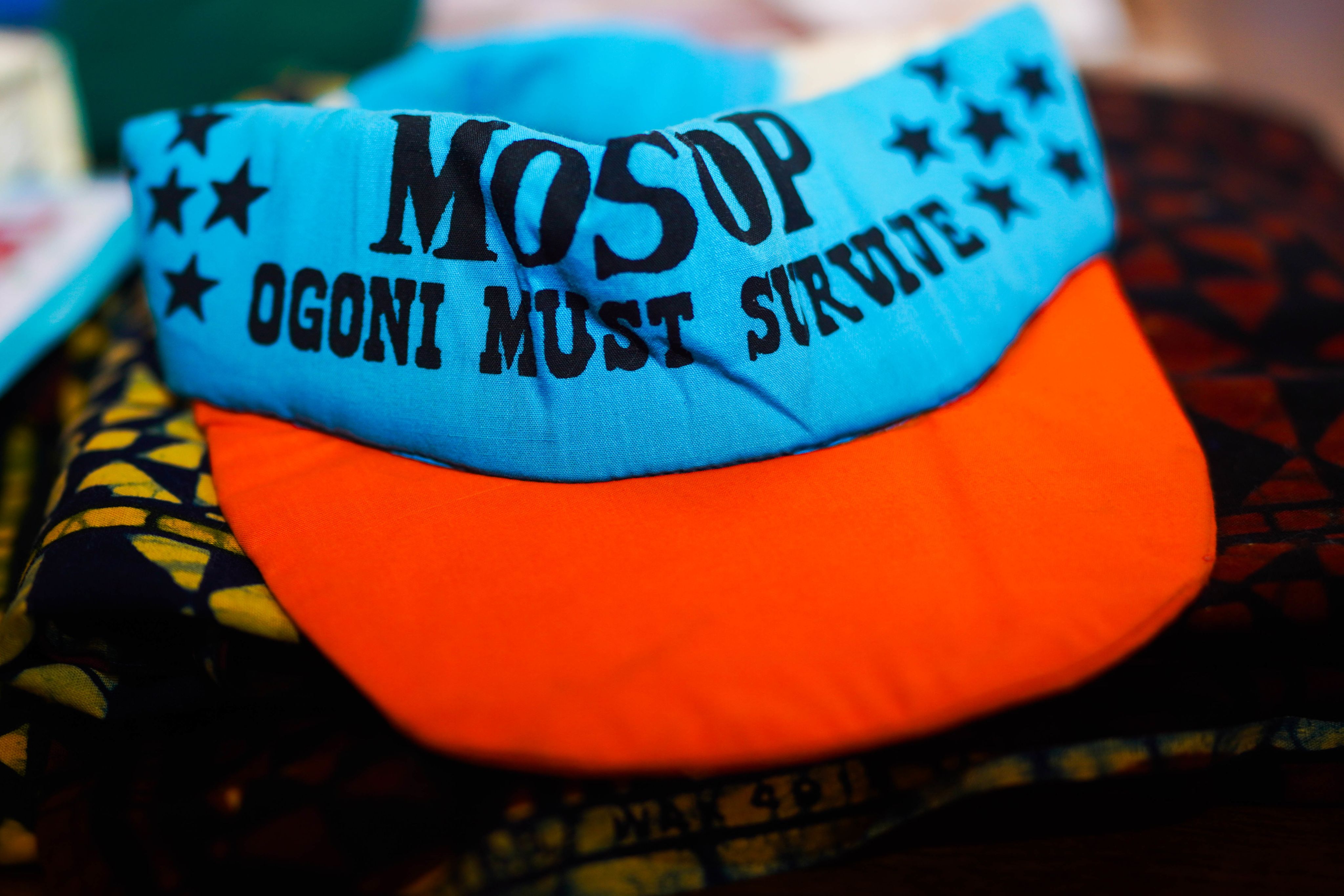 A bright blue visor with navy blue letters that read "MOSOP Ogoni Must Survive" and groups of navy blue stars. The visor has a red brim.