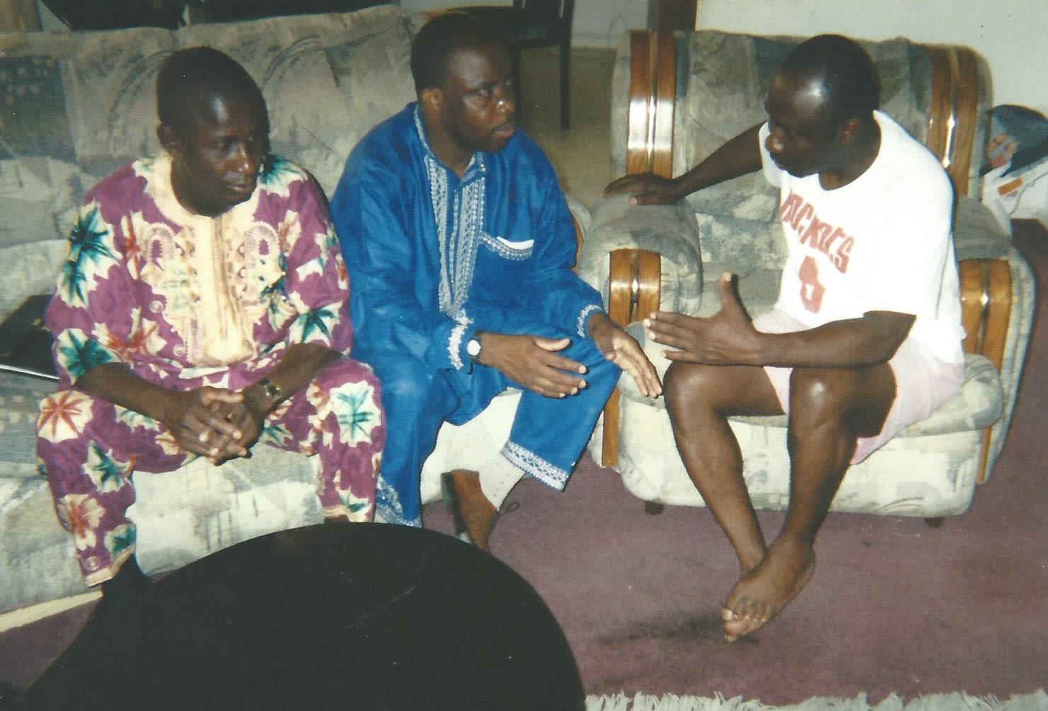 Two people in elaborate colorful dress sit on a couch next to a person in a white T-shirt.