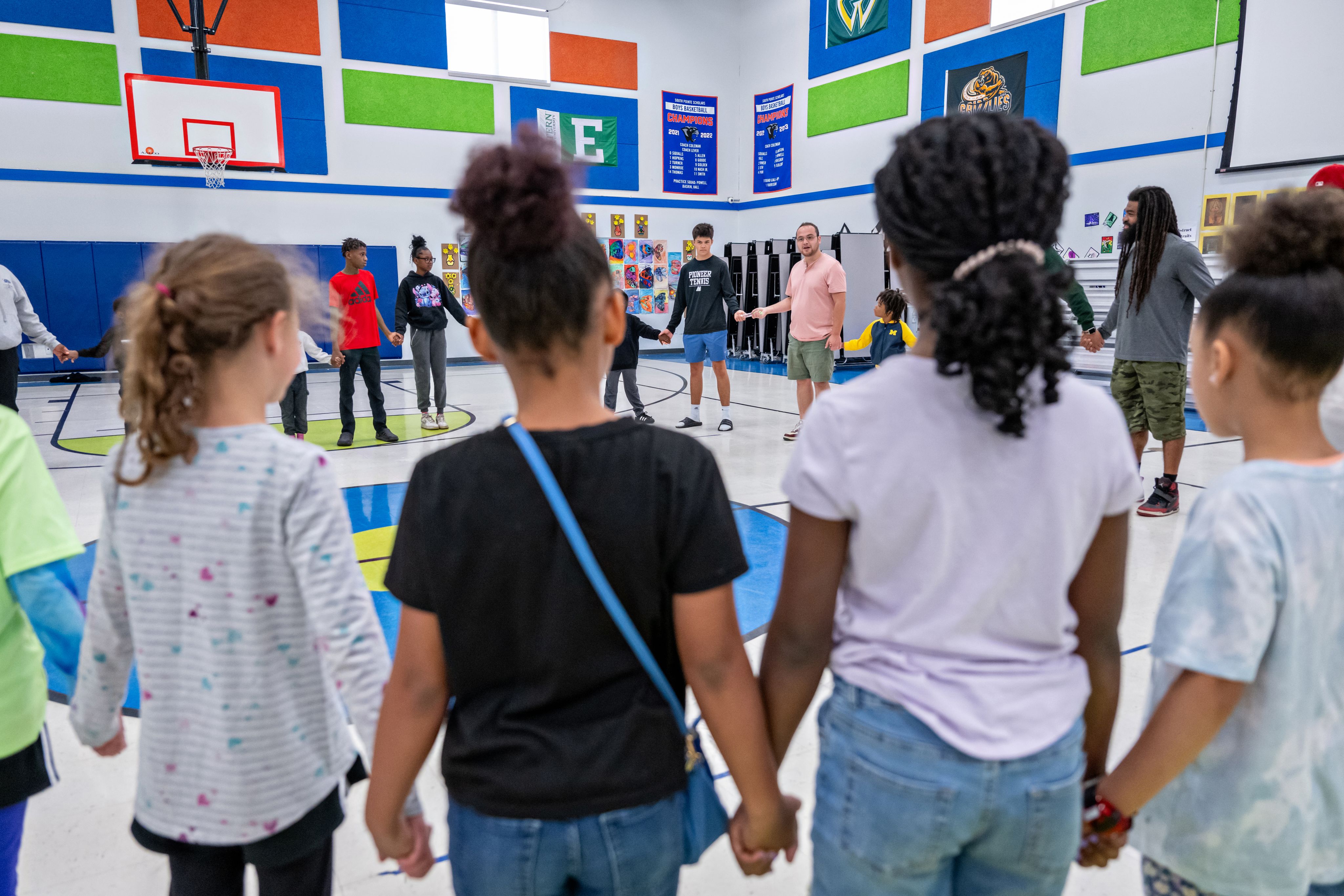 Children and adults hold hands in a circle in an elementary school gym.