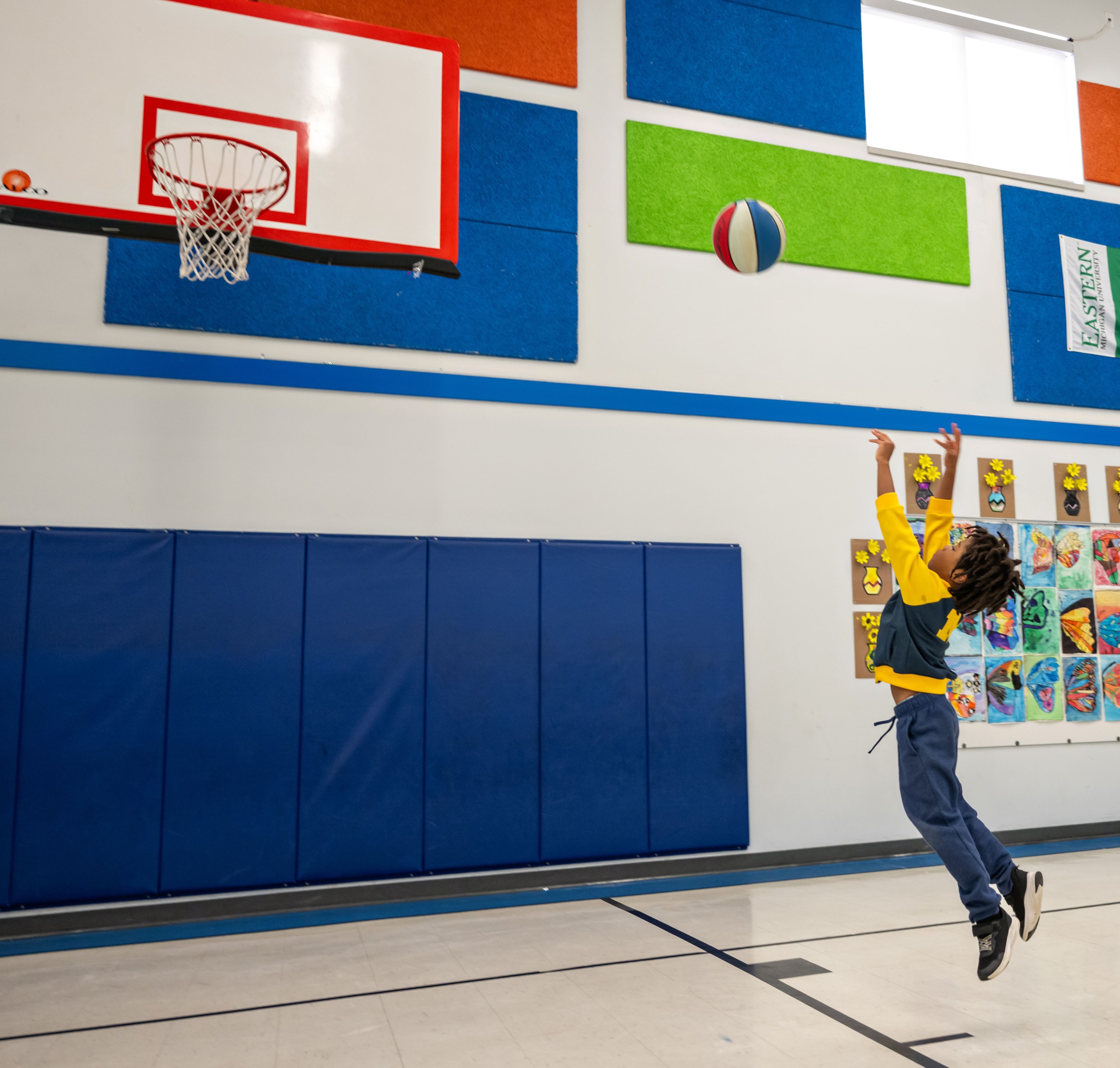 A kid in a U-M sweatshirt and navy blue sweatpants throws a basketball up toward a hoop in an elementary school gym with brightly colored squares on the walls.
